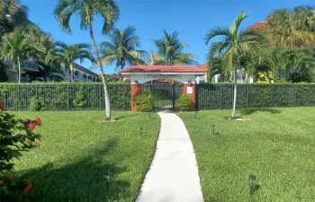 Gated entry featuring a terracotta-tiled archway, manicured lawn, perimeter fencing, and mature palm landscaping