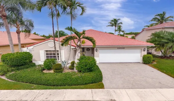 Single-story residence featuring a terracotta-tiled roof, paver driveway, attached two-car garage, and established landscaping with mature palm trees