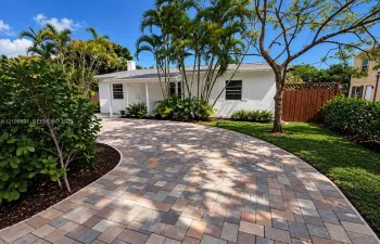 Single-story exterior featuring a multi-tone paver driveway, white stucco finish, front covered porch, and mature palm landscaping