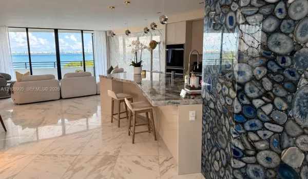 Polished stone flooring, expansive floor-to-ceiling windows with water scenery, custom waterfall-edge kitchen island, decorative blue agate stone accent wall, and built-in stainless steel appliances