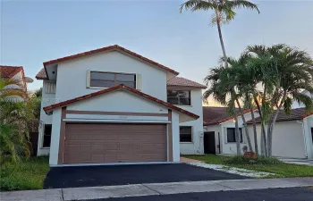 Two-story exterior featuring a light-toned stucco façade, terracotta-style roof tiles, an attached garage with a sectional door, and mature palm trees