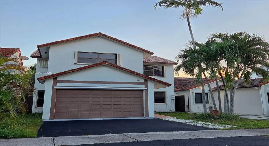 Two-story exterior featuring a light-toned stucco façade, terracotta-style roof tiles, an attached garage with a sectional door, and mature palm trees