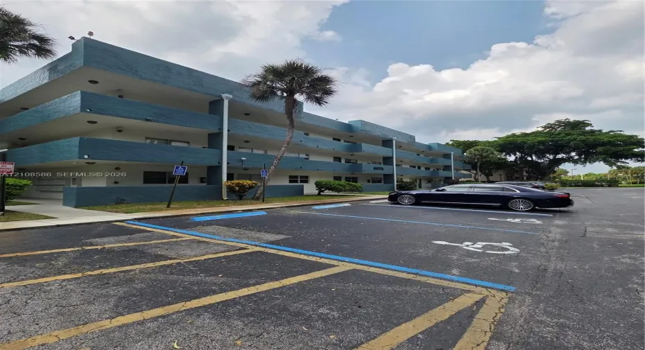 Three-story multi-unit structure featuring a textured blue facade, recessed balconies, and paved surface parking