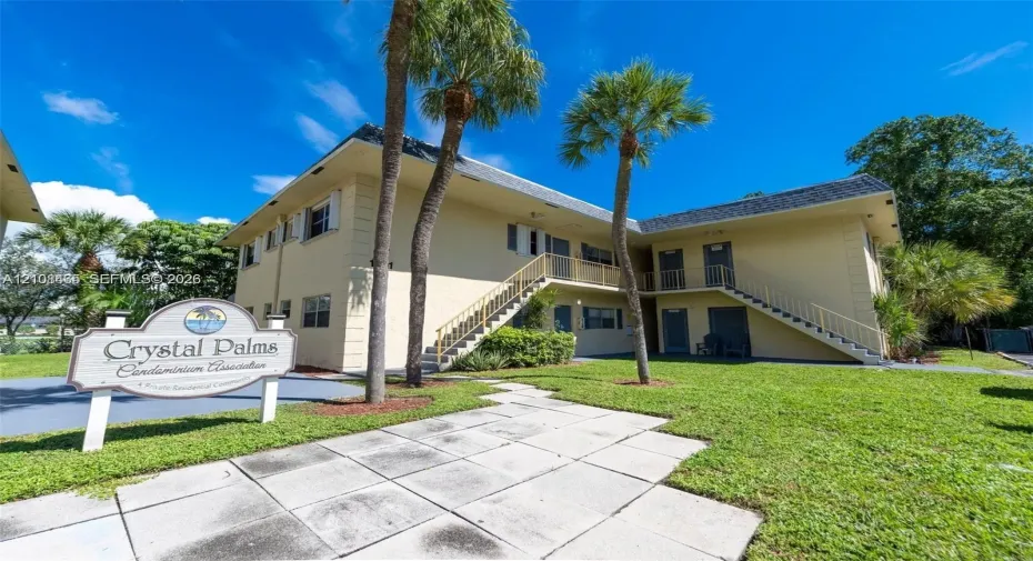 Two-story exterior featuring a light-toned facade, metal staircase railings, manicured lawn, and mature palm trees