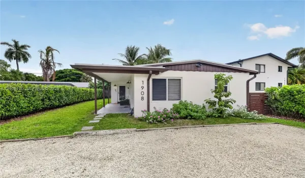 Single-story exterior featuring a covered front porch, light-colored stucco finish, stone-surfaced driveway, and mature perimeter hedging