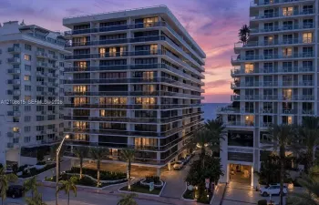 High-rise building exterior featuring glass-railed balconies, a paved porte-cochère entry, ground-level landscaping, and palm trees