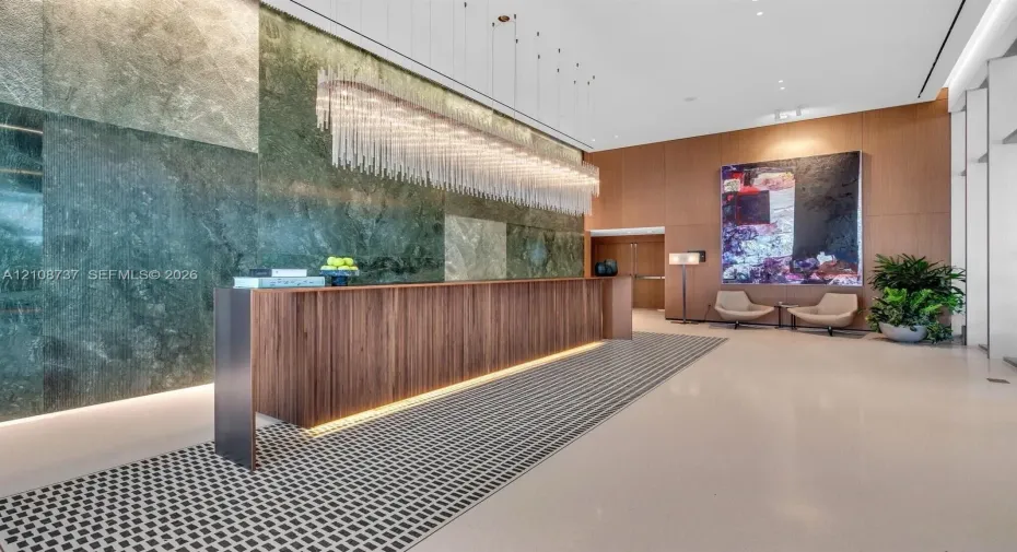 Grand lobby featuring a floor-to-ceiling polished stone accent wall, custom wood-finish reception desk with integrated lighting, elongated linear crystal chandelier, and large-format tile flooring