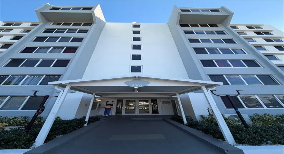 Multi-story concrete exterior featuring a covered entryway with a central pedestrian path, dual-wing facade architecture, and uniform window placement