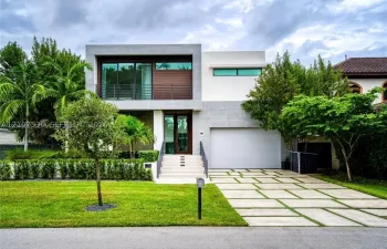 Modern architectural exterior featuring smooth stucco siding, wood-accented panels, a second-story balcony with horizontal metal railing, and a geometric concrete paver driveway