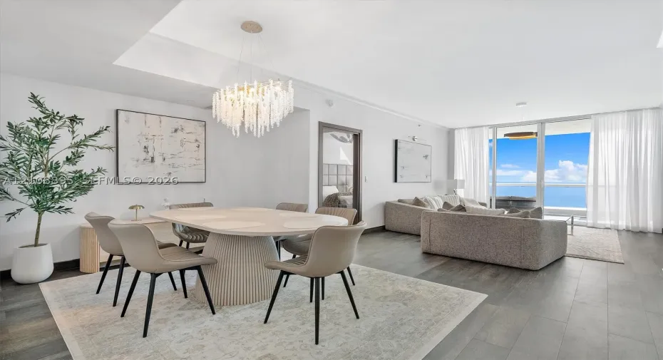 Open-concept living space featuring wood-finish flooring, a contemporary multi-tiered crystal chandelier, recessed ceiling lighting, and floor-to-ceiling glass paneling with an ocean backdrop