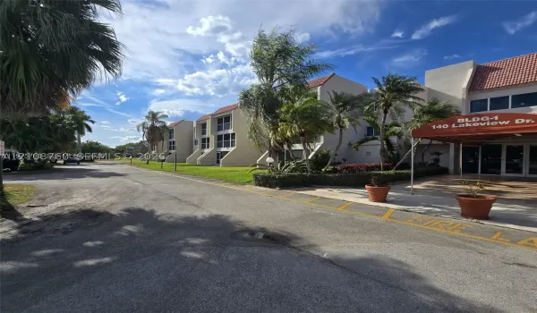 Multi-story exterior featuring a light-toned stucco façade, terra-cotta-hued roof tiles, and ground-level covered entryway with a red fabric awning, complemented by landscaped perimeter gardens and mature palms