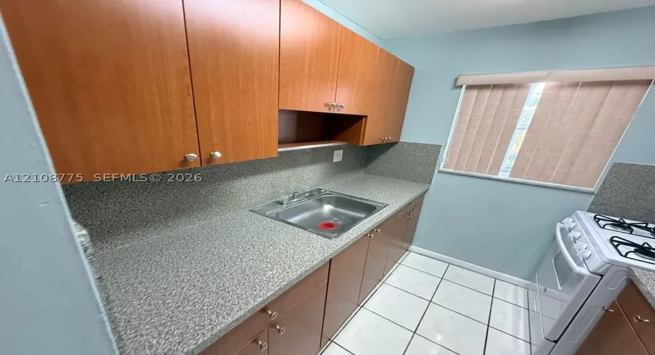Kitchen featuring wood-finish cabinetry, solid-surface countertops, integrated backsplash, stainless steel drop-in sink, and ceramic tile flooring