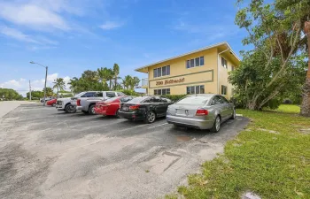 Multi-story building exterior featuring a low-pitch roofline, stucco-finish facade, designated surface parking, and mature tropical landscaping