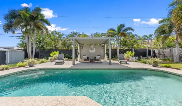 In-ground swimming pool with stone-texture decking, shaded pergola, mature palms, and perimeter privacy fencing
