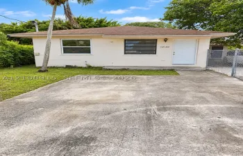 Single-story exterior featuring a hip roof, stucco facade, concrete driveway, and chain-link fencing