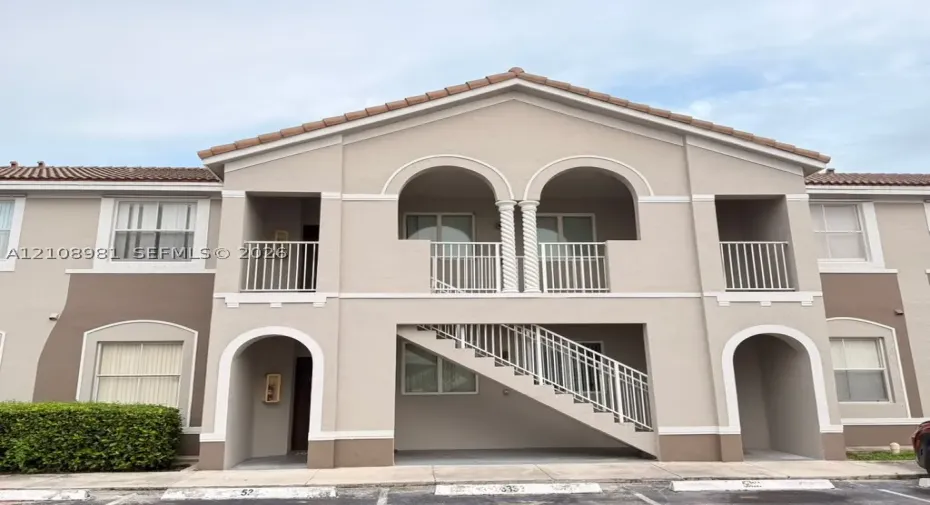 Exterior building facade featuring arched architectural details, twin columns on the upper balcony, clay-tile roofing, and metal stair railings