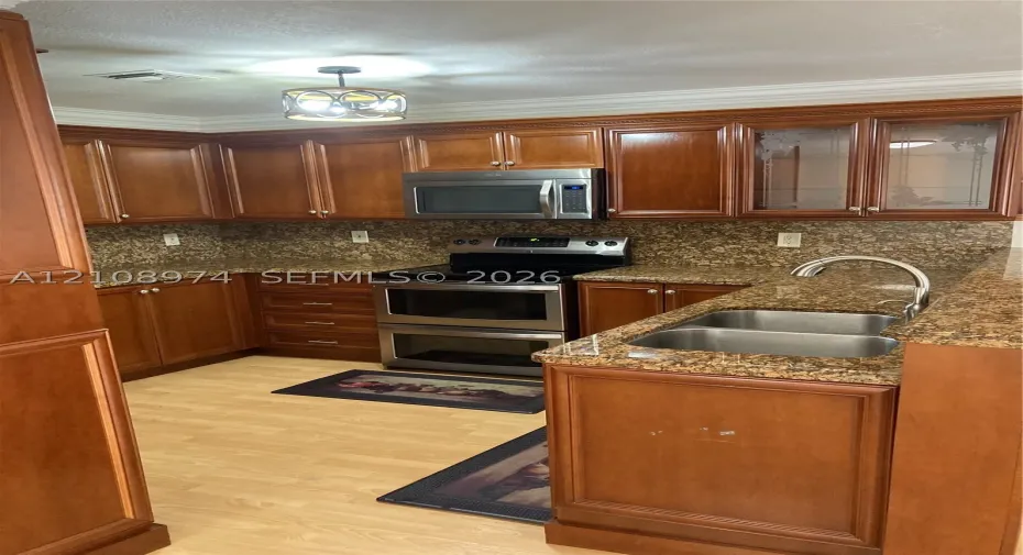 Kitchen featuring stained wood cabinetry, stone-patterned countertops, stainless steel appliances, a dual-basin undermount sink, and crown molding