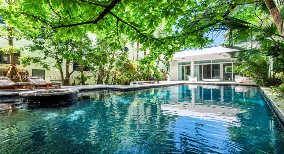 In-ground swimming pool featuring stone coping and a circular integrated spa, surrounded by mature trees, lush tropical landscaping, and expansive stone decking