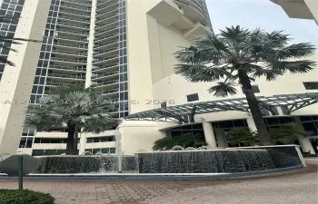 High-rise exterior featuring curved glass balconies, a circular fountain with tiered stone masonry, a metal-framed porte-cochere, and mature palm landscaping