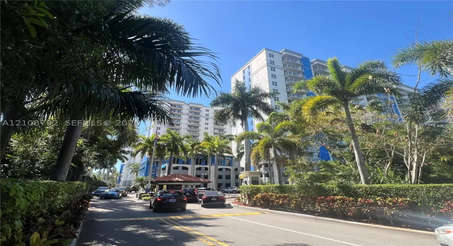 High-rise condominium building featuring tiered balconies, gated entry structure with a tile roof, mature palm trees, and manicured perimeter landscaping