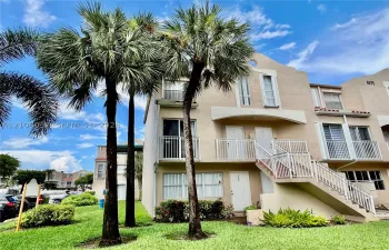 Multi-story stucco exterior featuring white metal balcony railings, a staircase with matching railing, and mature palm trees