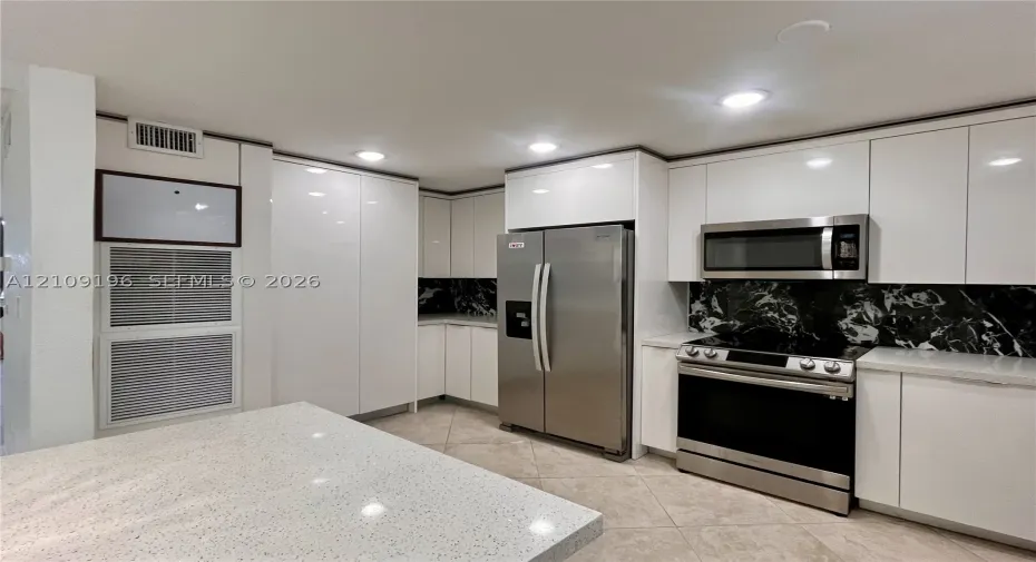 Kitchen featuring sleek white flat-panel cabinetry, light-toned stone-finish countertops, stainless steel appliances, and a high-contrast dark stone backsplash