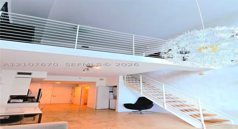 Double-height ceilings, mezzanine level with horizontal metal railing, custom dandelion-style pendant light, open staircase with wood-finish treads, and stone tile flooring