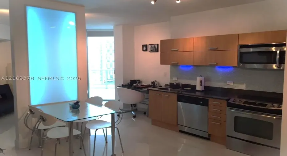 Kitchen featuring wood-finish cabinetry, stainless steel appliances, dark stone-surface countertops, glass tile backsplash with integrated under-cabinet lighting, and an illuminated wall panel