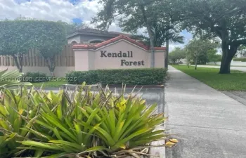 Stucco monument signage with tile detailing, manicured shrubbery, mature trees, and concrete sidewalk