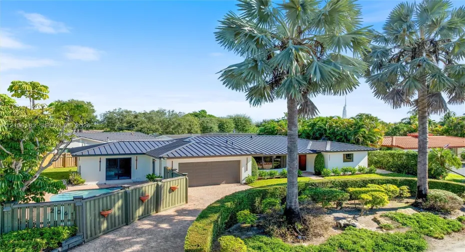 Single-story residence featuring a standing seam metal roof, attached multi-car garage, gated entry, integrated swimming pool, and mature palm landscaping