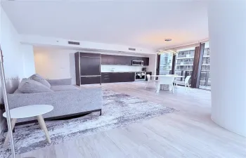 Open-concept living space featuring wood-finish flooring, dark cabinetry, stainless steel appliances, floor-to-ceiling glass, and a modern sculptural pendant light fixture