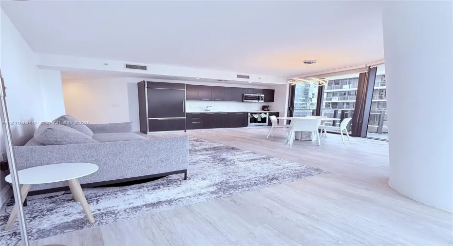 Open-concept living space featuring wood-finish flooring, dark cabinetry, stainless steel appliances, floor-to-ceiling glass, and a modern sculptural pendant light fixture