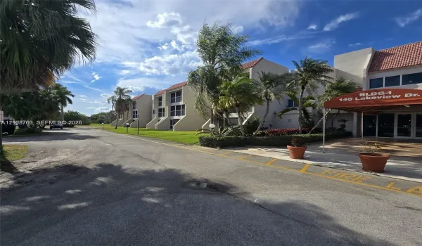 Multi-story exterior featuring a light-toned stucco façade, terra-cotta-hued roof tiles, and ground-level covered entryway with a red fabric awning, complemented by landscaped perimeter gardens and mature palms