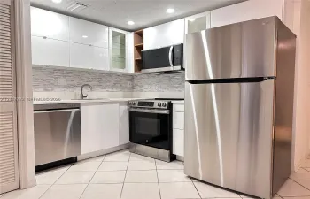 Kitchen featuring high-gloss white cabinetry, stainless steel appliances, a multi-tonal tile backsplash, integrated microwave, and neutral tile flooring