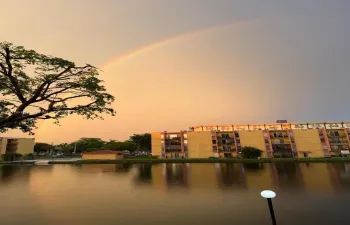 Multi-story building exterior featuring balconies and water-front access overlooking a pond with mature trees and a rainbow backdrop
