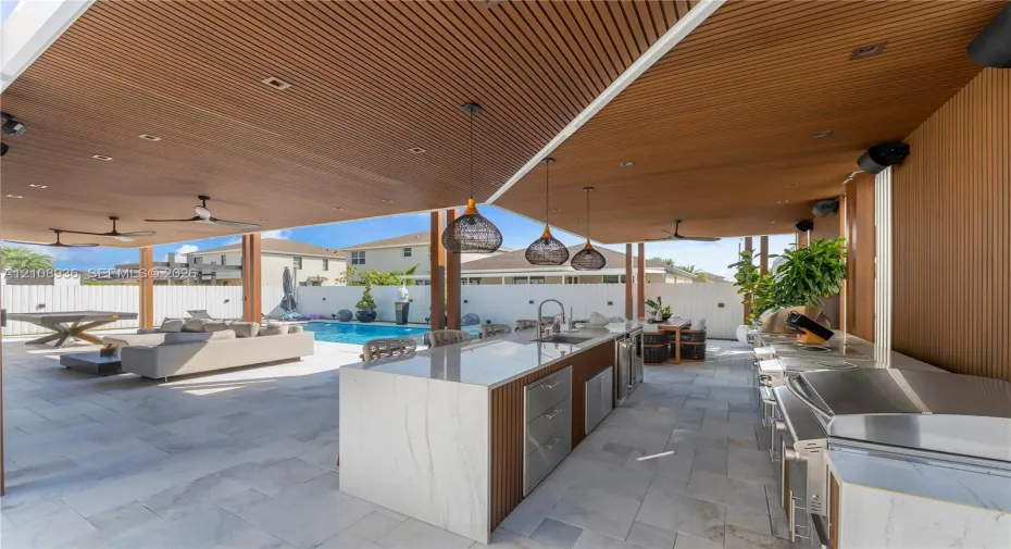 Covered outdoor kitchen featuring stone-finish counters, stainless steel built-in grill, and wood-slat ceiling accents, overlooking a private swimming pool with stone-tile patio flooring