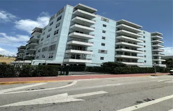 Multi-story residential building featuring a light blue stucco exterior, stacked private balconies with black metal railings, and a gated ground-level parking entrance