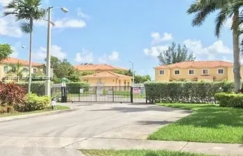 Gated entrance featuring a metal security gate, concrete driveway, and mature perimeter landscaping