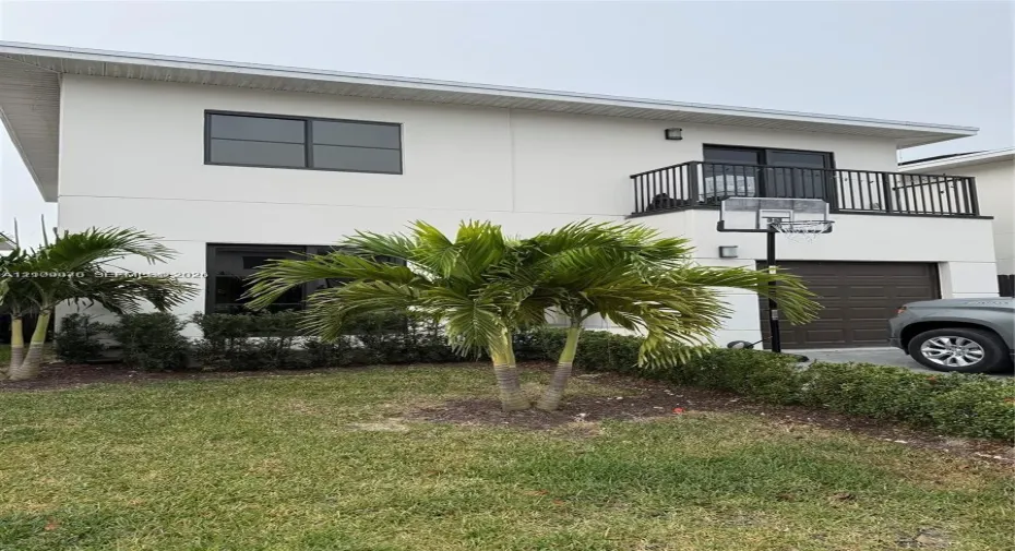 Two-story contemporary exterior featuring a smooth stucco facade, metal-framed windows, a private balcony with black metal railing, and an attached single-car garage with a paneled door
