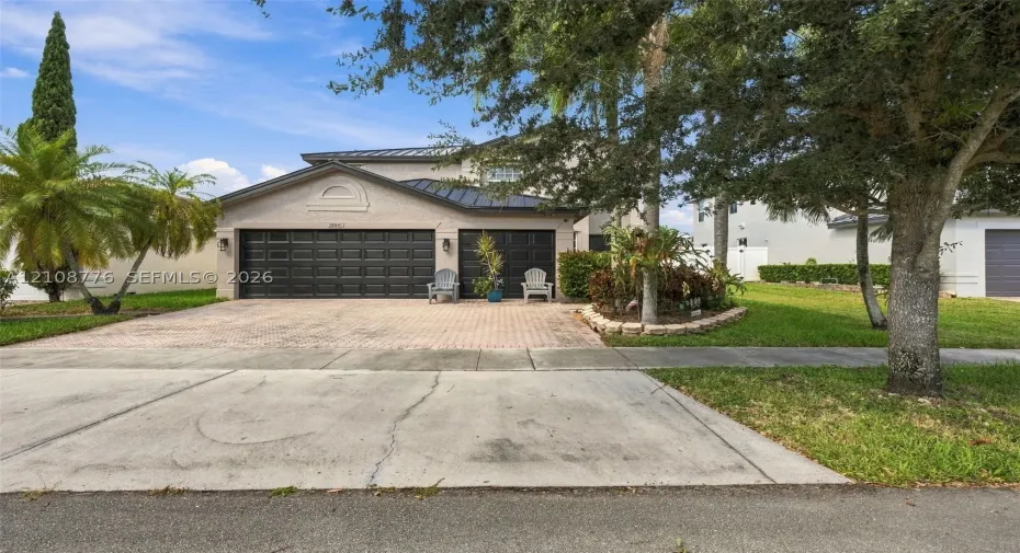 Multi-level exterior featuring a metal roof, paver driveway, three-car garage with dark-finish doors, and landscaped garden beds