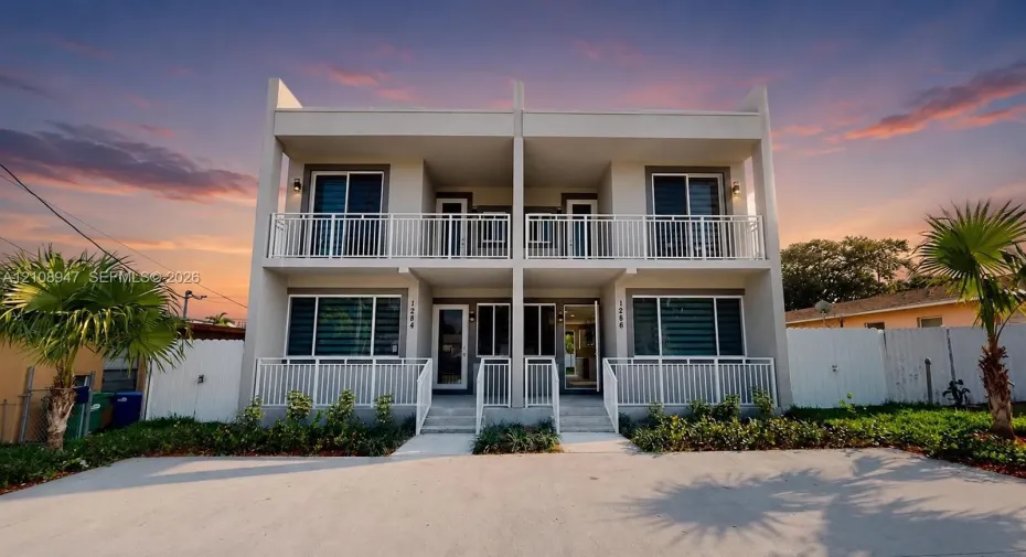 Two-story stucco exterior featuring dual-level white metal railed balconies, concrete hardscaping, and integrated exterior lighting