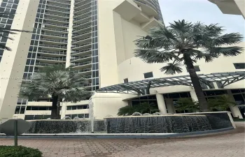 High-rise exterior featuring curved glass balconies, a circular fountain with tiered stone masonry, a metal-framed porte-cochere, and mature palm landscaping