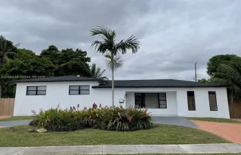 Single-story exterior featuring white stucco finish, dark-toned roof shingles, large multi-pane windows, and a mature palm tree