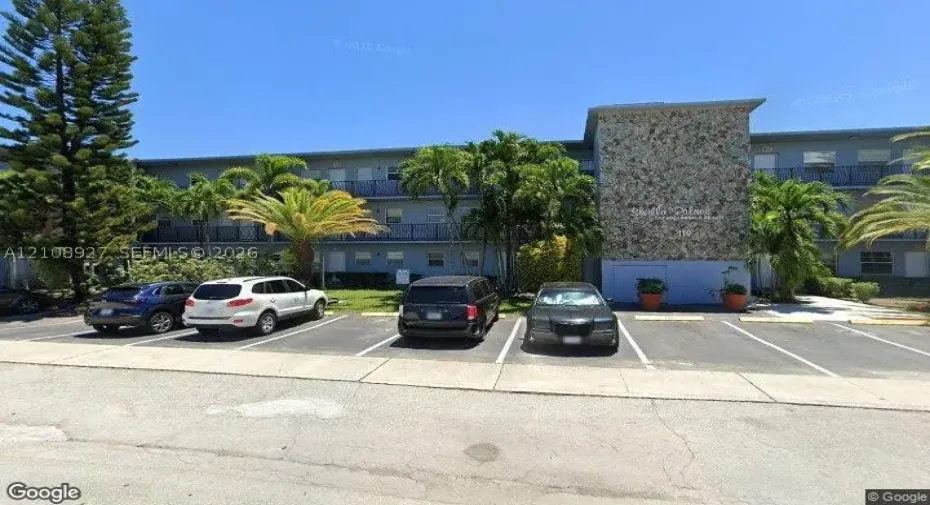Multi-story exterior featuring a natural stone facade, dedicated surface parking, and mature palm landscaping