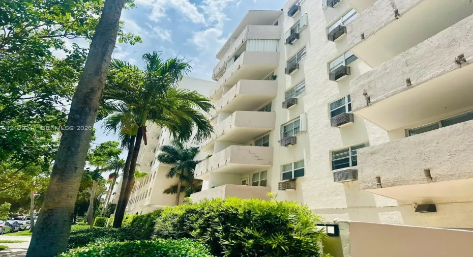 Multi-story residential building exterior featuring a stucco facade, private balconies, and mature palm landscaping