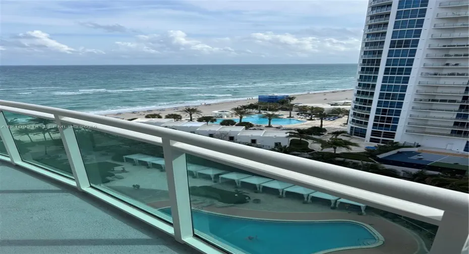 Private balcony featuring glass railings, overlooking a swimming pool, cabanas, and oceanfront shoreline