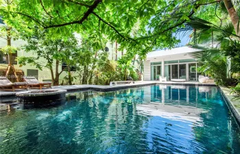 In-ground swimming pool featuring stone coping and a circular integrated spa, surrounded by mature trees, lush tropical landscaping, and expansive stone decking