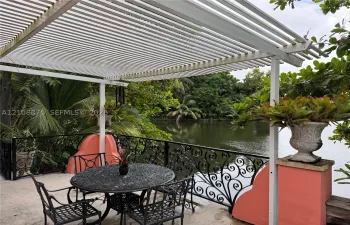 Outdoor patio featuring a white louvered pergola, black wrought-iron railing, concrete flooring, and decorative masonry pillars