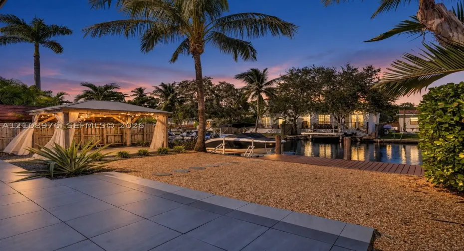 Waterfront property featuring a covered timber-framed gazebo, wooden dock, gravel landscaping, and large-format tile patio