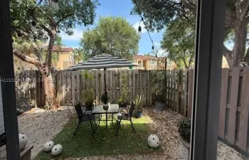 Private fenced outdoor area featuring a pebble ground covering, a designated patch of turf, mature trees, and overhead string lighting